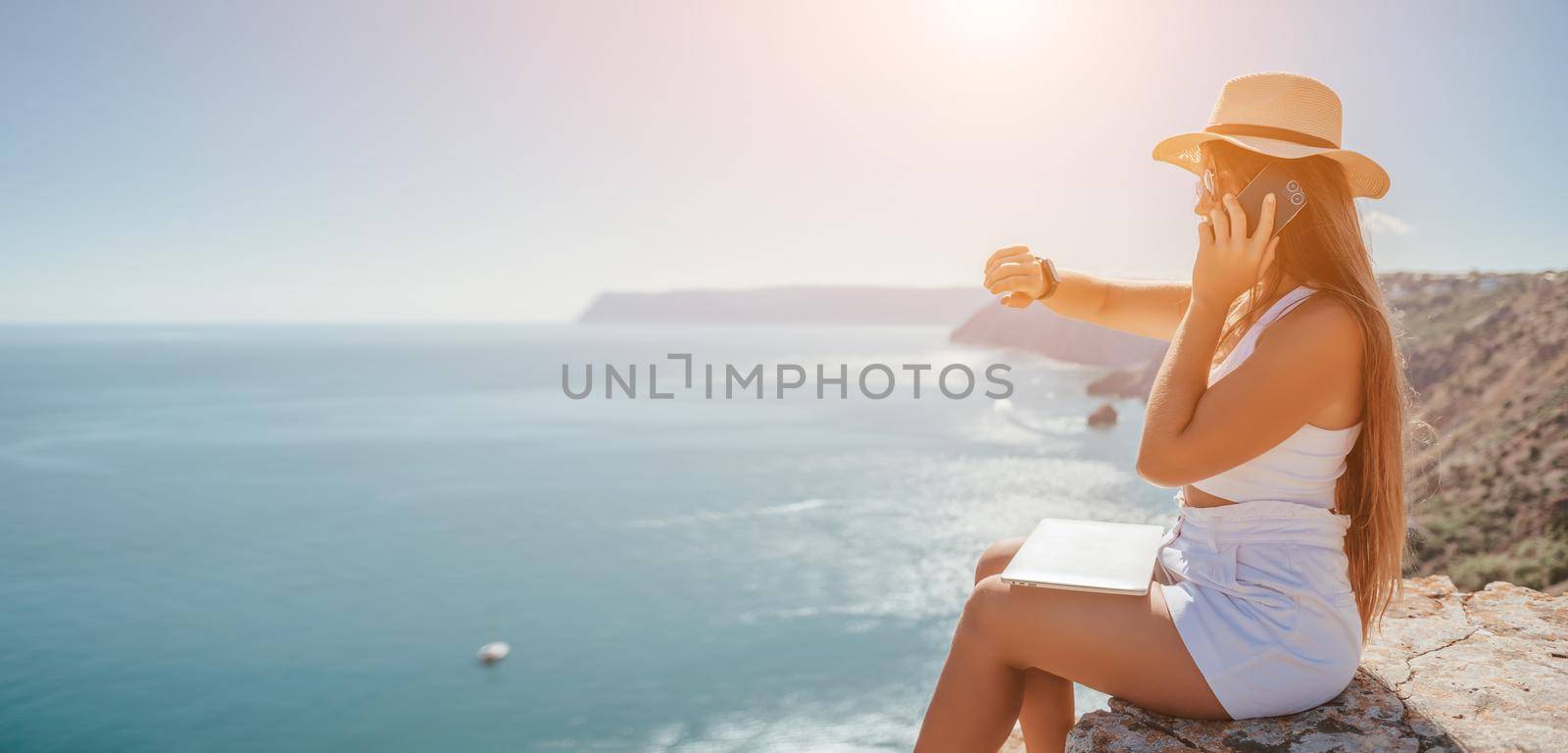 Successful business woman in yellow hat working on laptop by the sea. Pretty lady typing on computer at summer day outdoors. Freelance, travel and holidays concept.