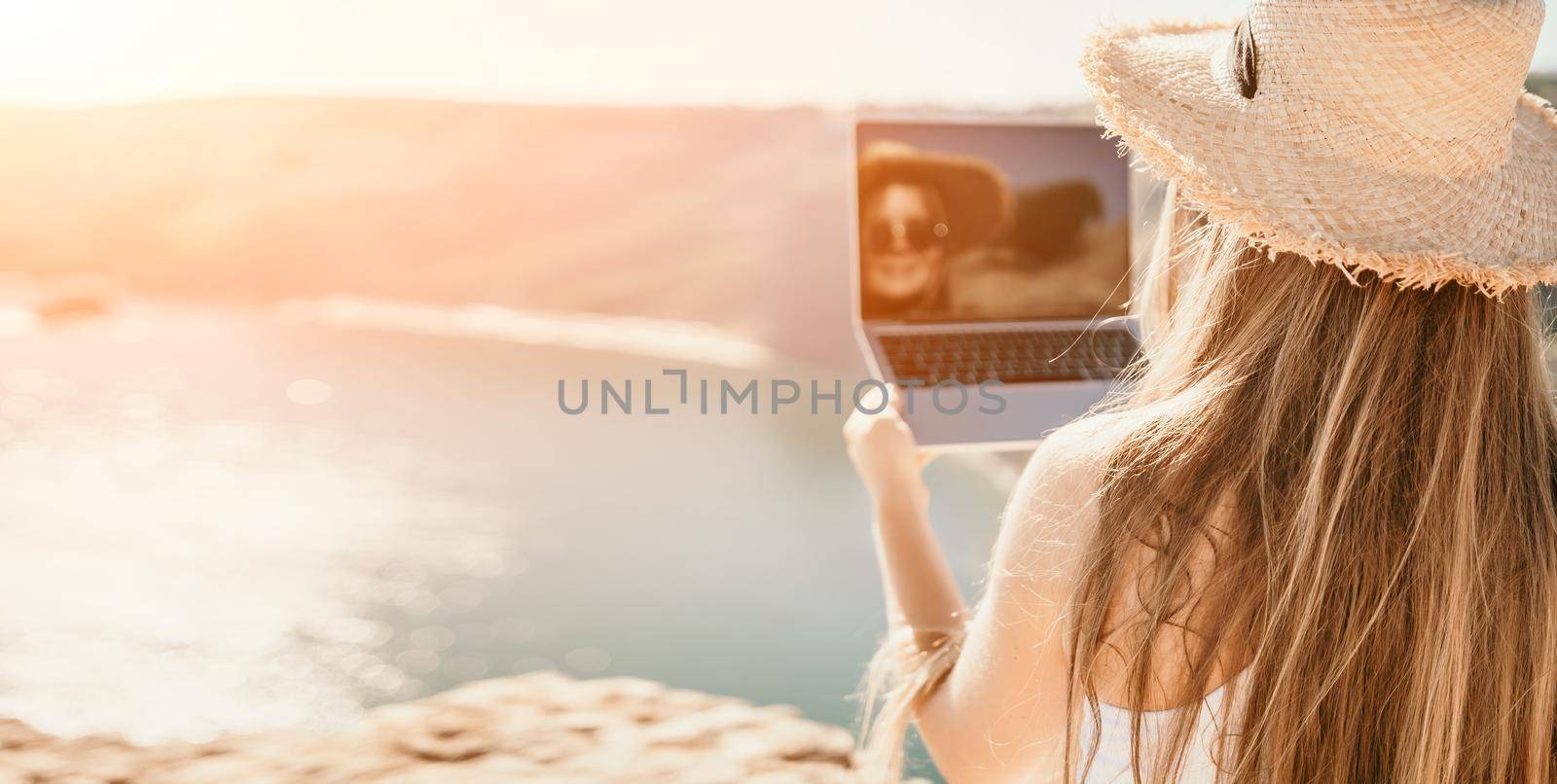Successful business woman in yellow hat working on laptop by the sea. Pretty lady typing on computer at summer day outdoors. Freelance, travel and holidays concept. Back view.