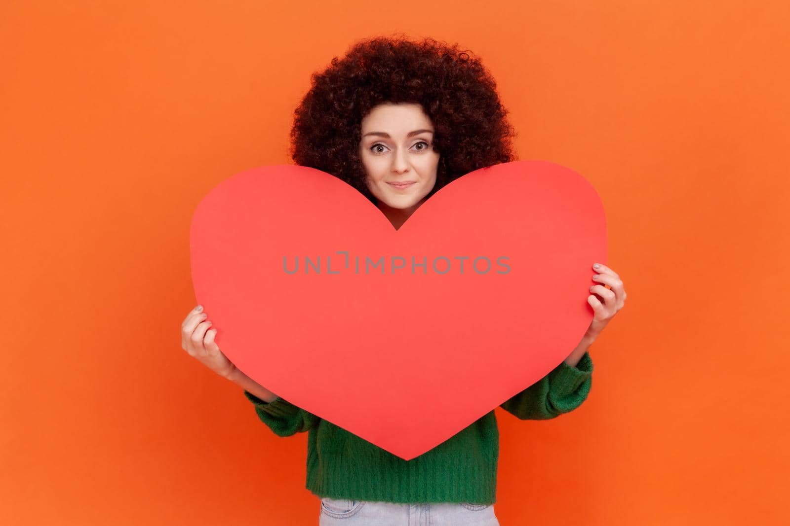 Positive kind woman with Afro hairstyle wearing green casual style sweater holding big red paper heart, looking at camera with friendly expression. Indoor studio shot isolated on orange background.