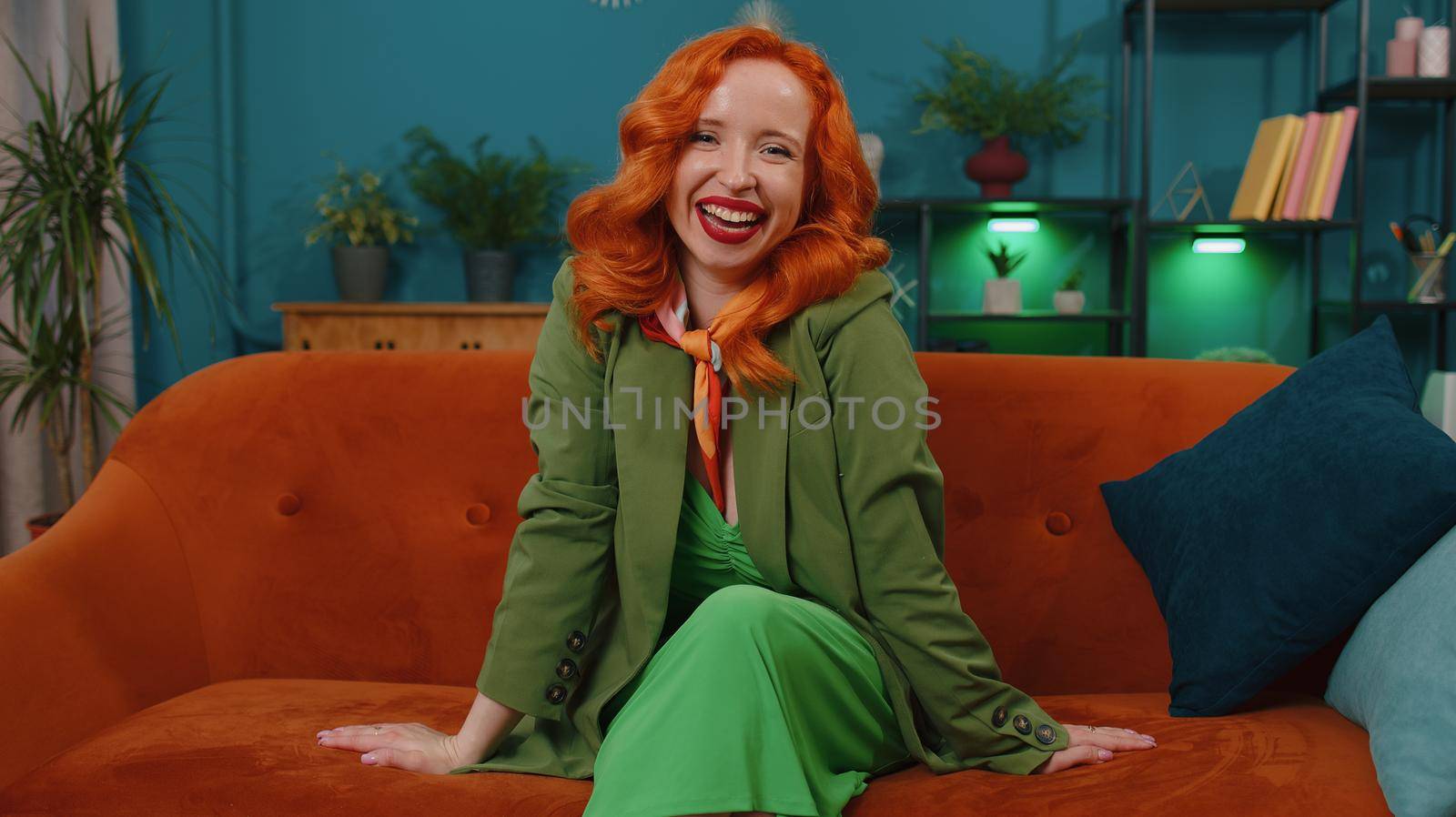 Redhead young woman in green dress laughing out loud after hearing ridiculous anecdote, funny joke, feeling carefree amused, positive people lifestyle at home. Ginger girl in room sitting on sofa