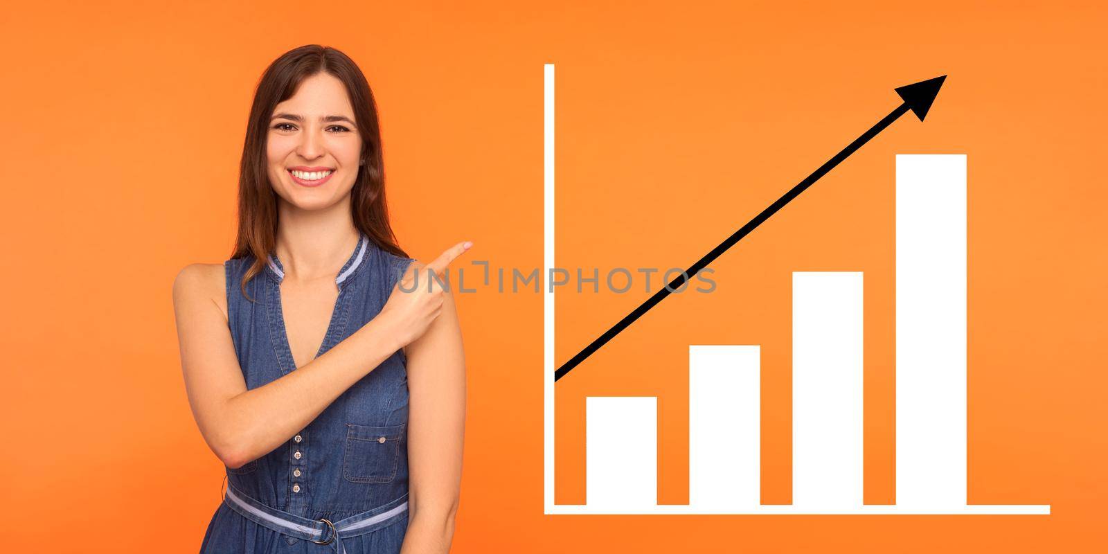 Portrait of happy joyful young woman standing, pointing aside and showing business growth graph. indoor studio shot isolated on orange background