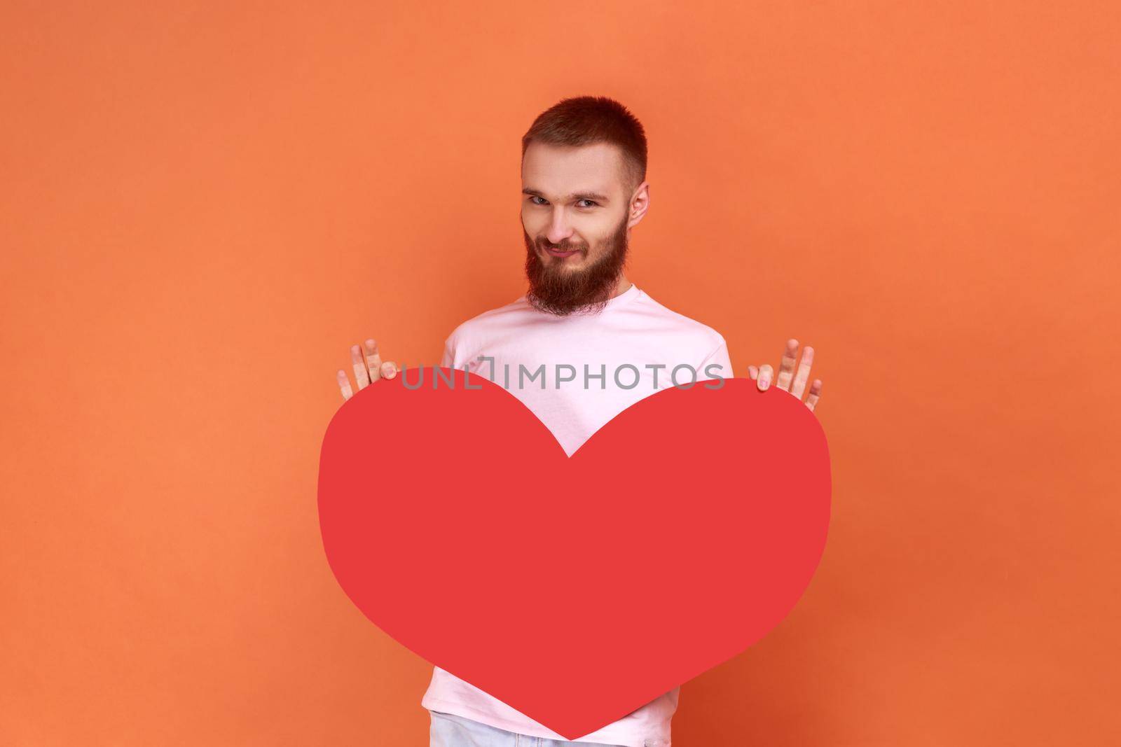 Portrait of positive optimistic young bearded man holding big red heart, expressing positive romantic emotions, wearing pink T-shirt. Indoor studio shot isolated on orange background.