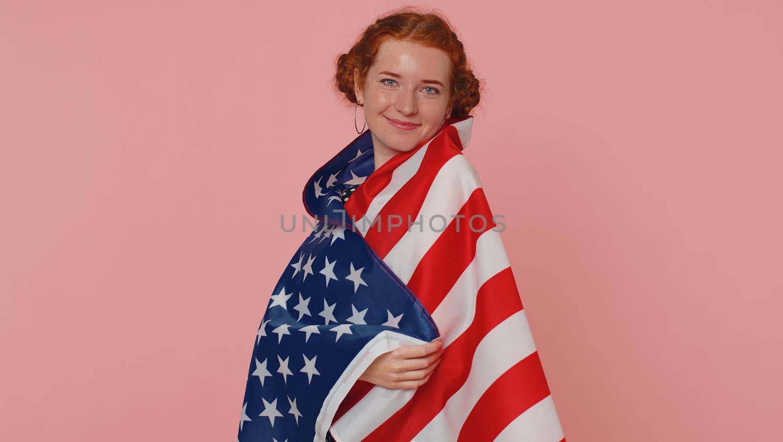 Young woman in polkadot dress waving and wrapping in American USA flag, celebrating, human rights and freedoms. Independence day. Ginger girl with freckles isolated alone on pink studio background