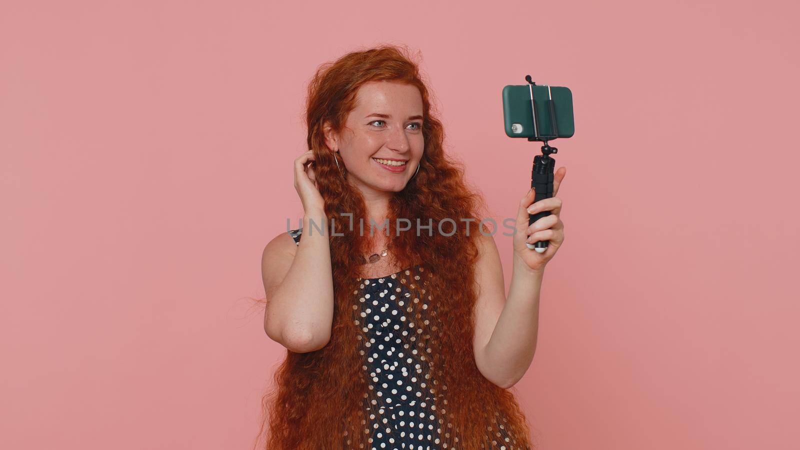 Redhead woman in dress traveler blogger taking selfie on mobile phone selfie stick, communicating video call online with subscribers. Young ginger girl isolated alone on pink studio background