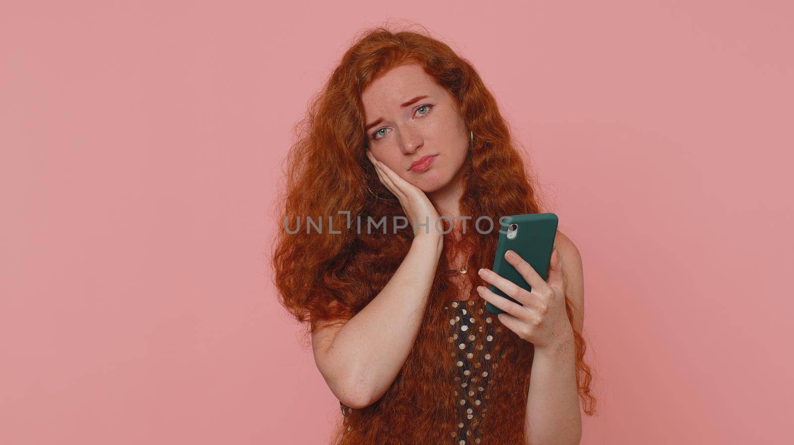 Unhappy sad young redhead woman use smartphone typing browsing, loses becoming surprised sudden lottery results, bad fortune, loss, unlucky news. Ginger girl with freckles on pink studio background