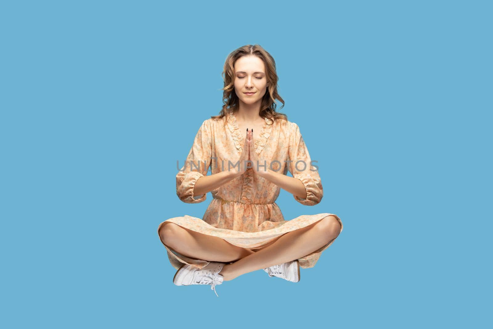 Hovering in air. Peaceful calm relaxed girl yellow dress levitating with prayer gesture, keeping eyes closed, meditating sitting in yoga position. indoor studio shot isolated on blue background