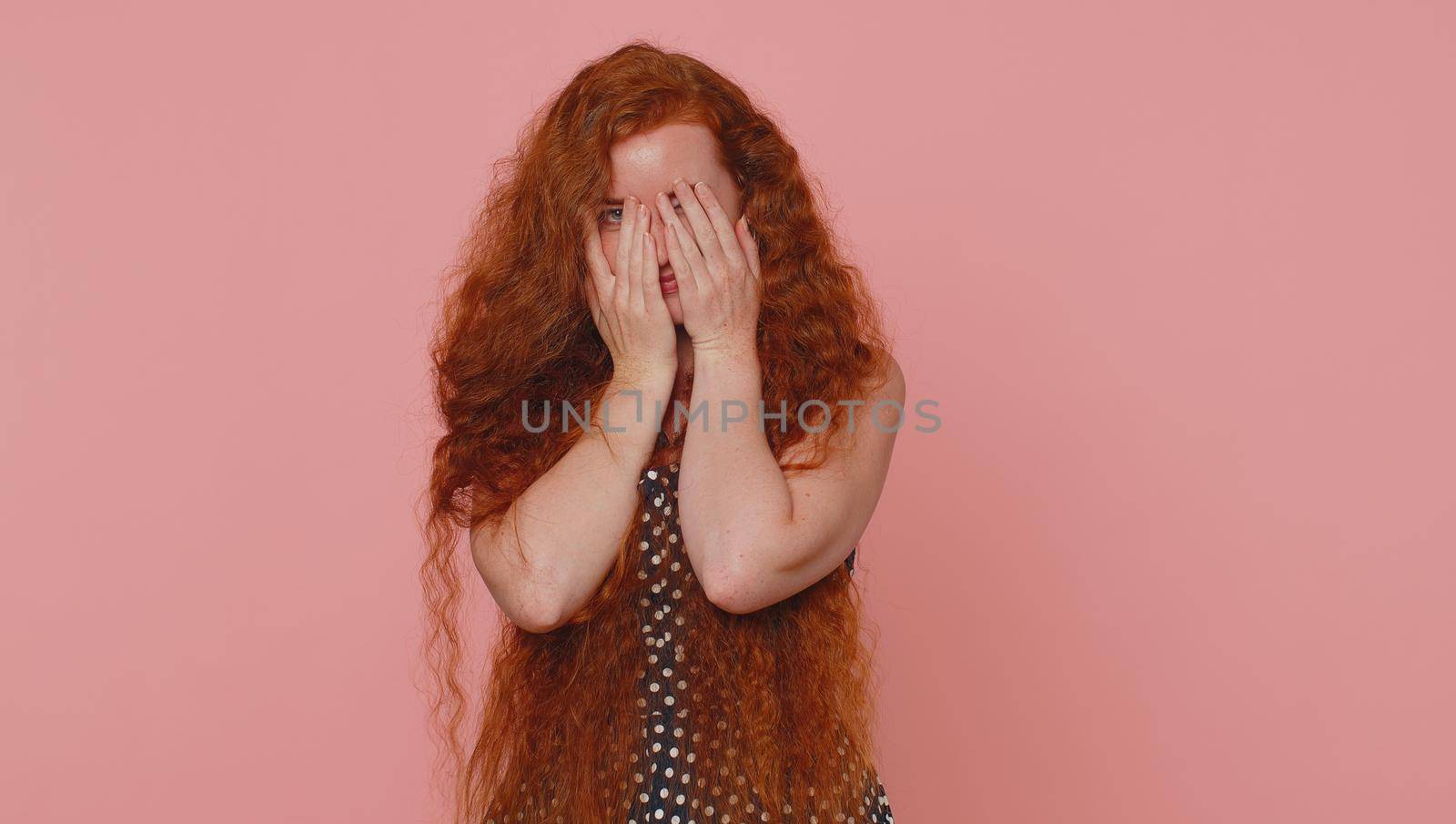 Nosy curious young redhead woman closing eyes with hand and spying through fingers, hiding and peeping, binocular gesture, exploring way, seeking something in distance. Ginger girl on pink background