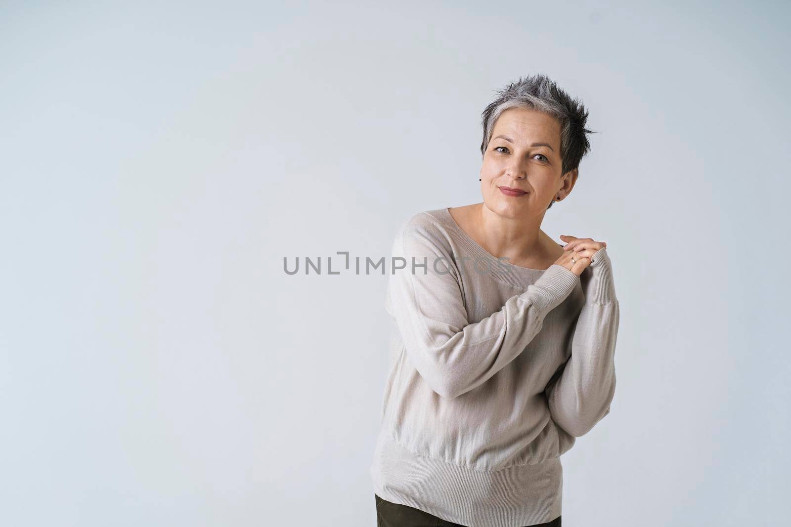 Smiling mature grey short hair woman posing with hands folded at her shoulder looking at camera wearing white blouse, copy space isolated on white background. Healthcare, aged beauty concept by LipikStockMedia