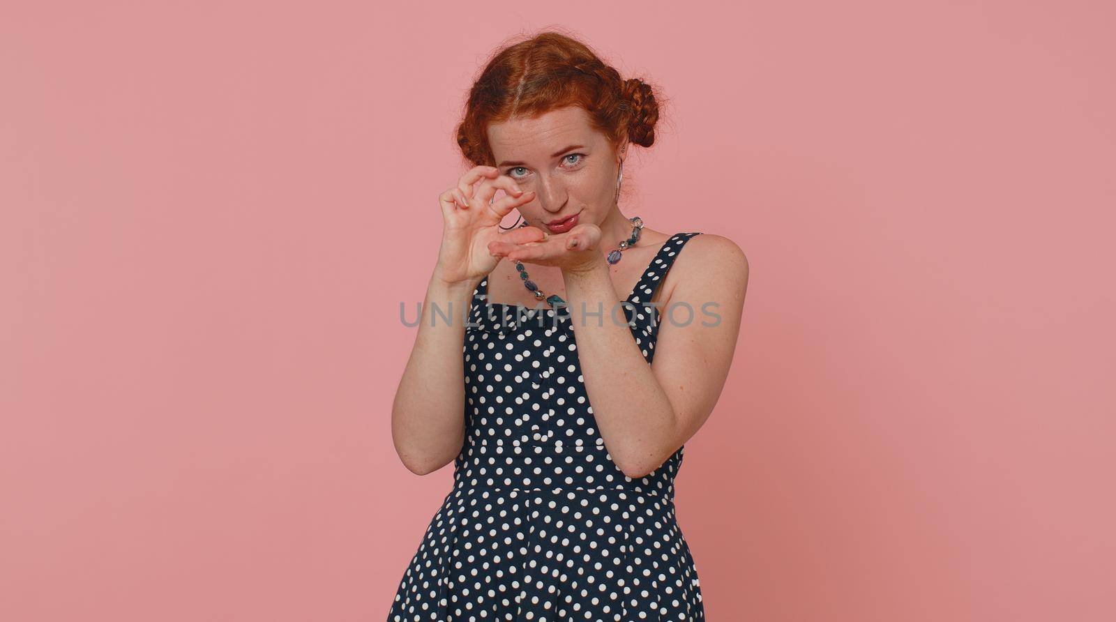 Need some more please give me. Woman showing a little bit gesture with sceptic smile, showing minimum sign, measuring small size asking for help. Ginger girl isolated alone on pink studio background