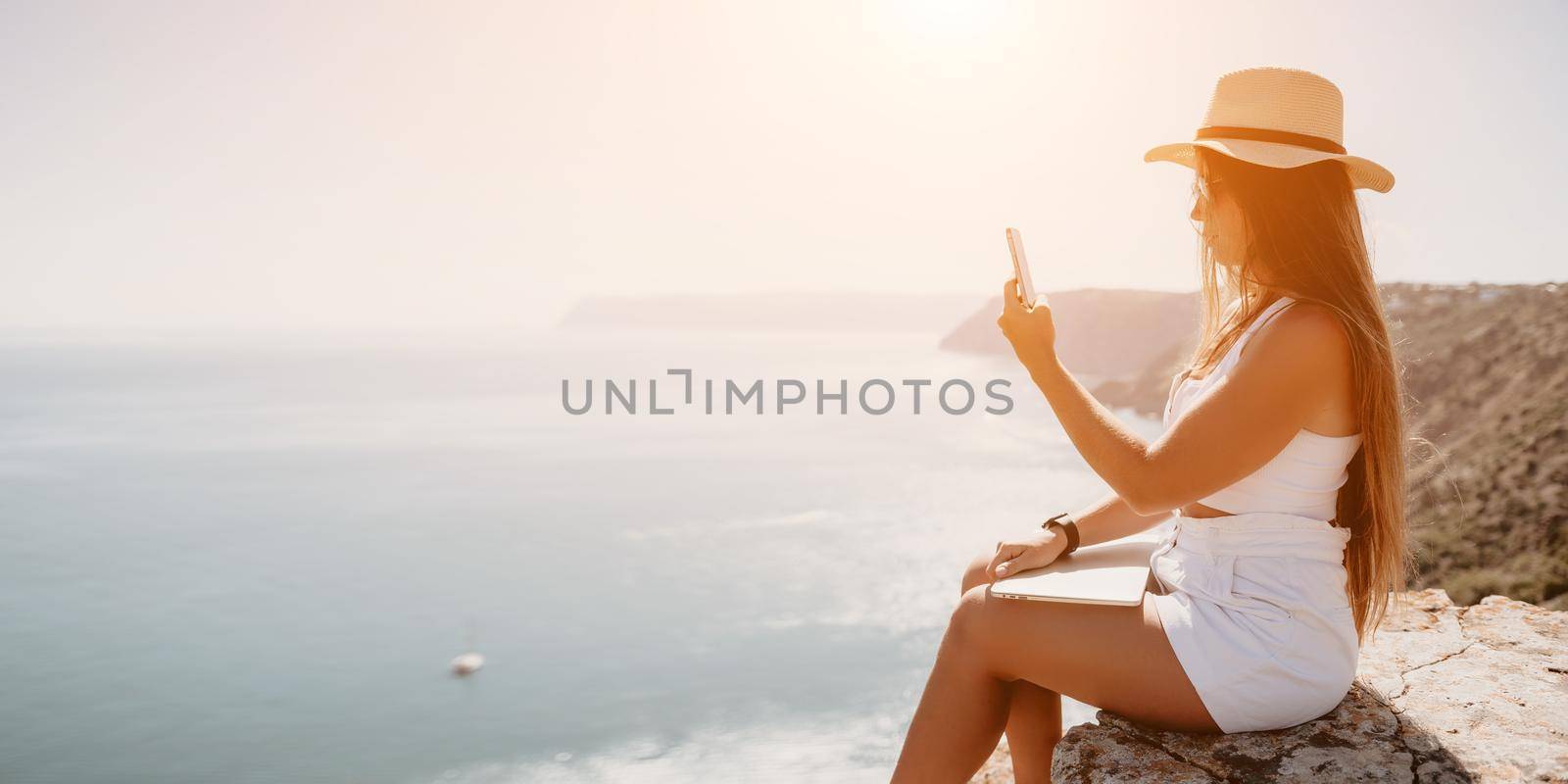 Successful business woman in yellow hat working on laptop by the sea. Pretty lady typing on computer at summer day outdoors. Freelance, travel and holidays concept.