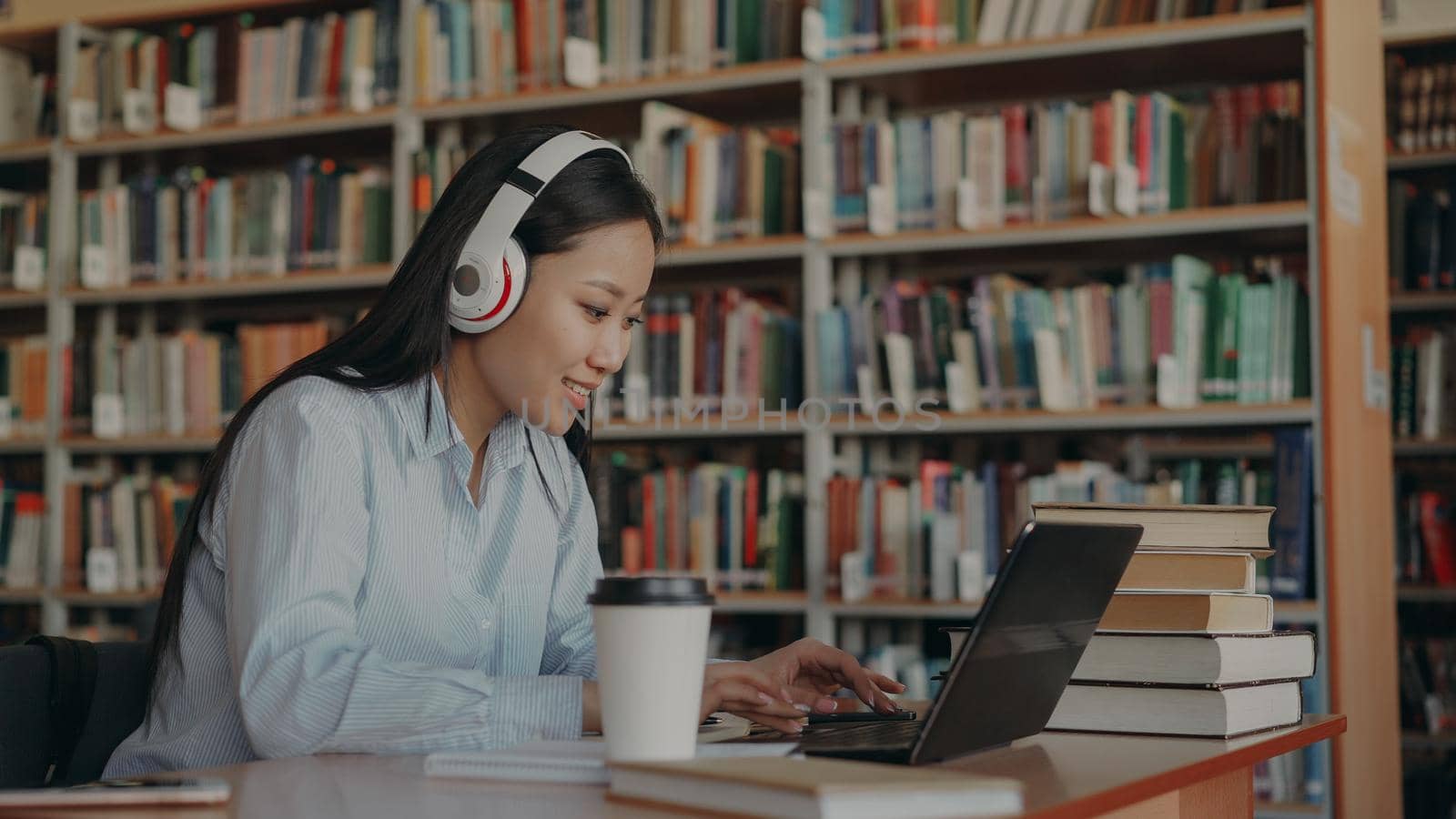 Beautiful asian female student with big headphones is sitting at desk in library working at school project.