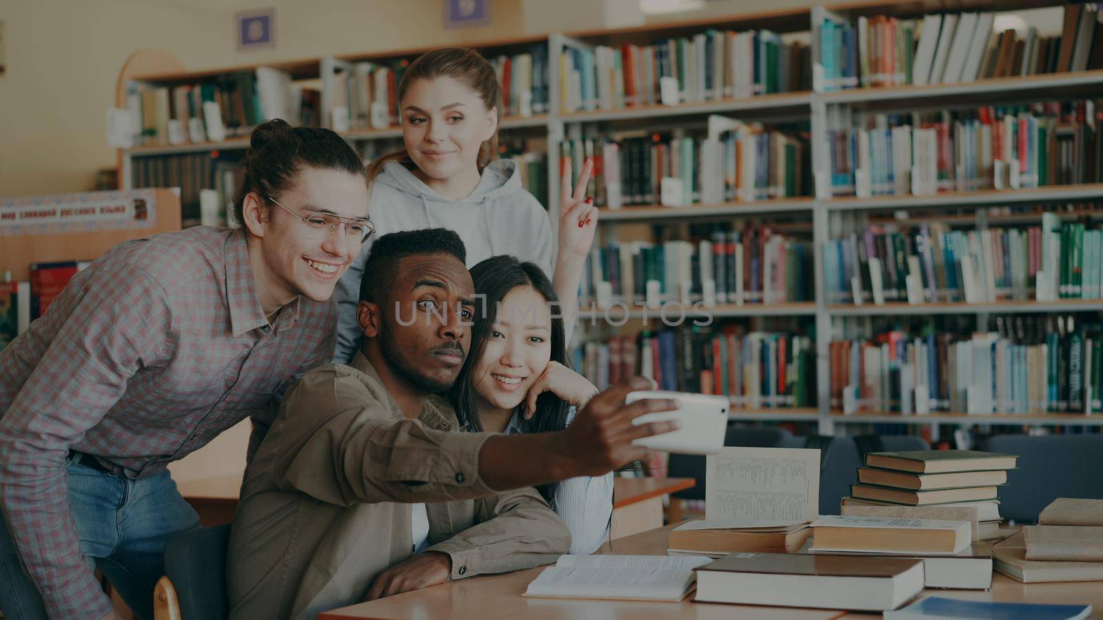 Group of international students have fun smiling and making selfie photos on smartphone camera at university library. Cheerful friends have rest while prepare for examination by silverkblack