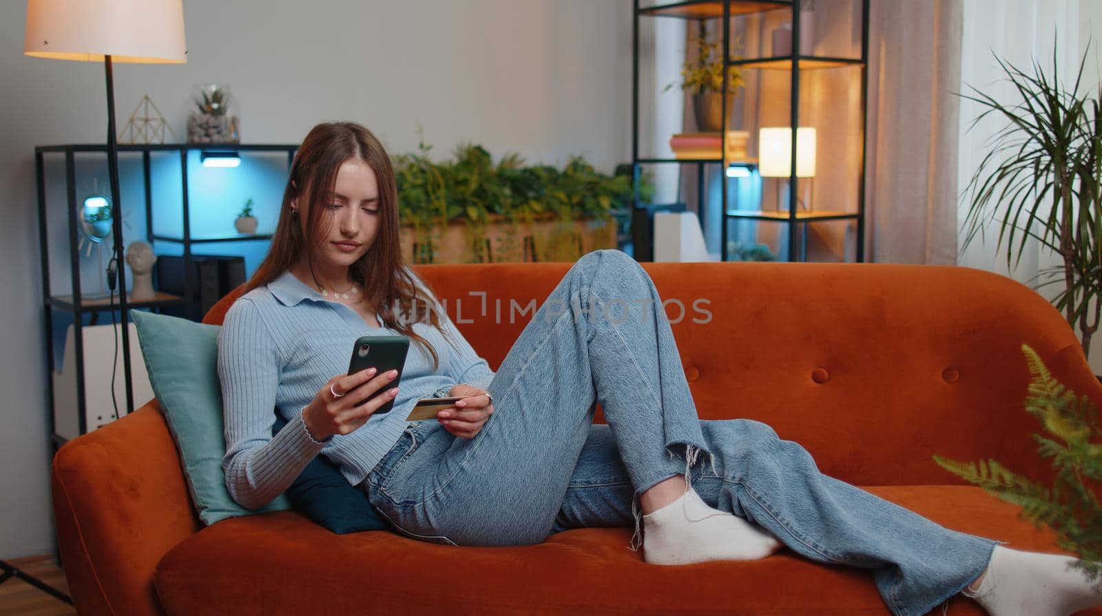 Young woman using credit bank card and smartphone while transferring money, purchases online shopping, order food delivery at modern home apartment indoors. Pretty girl in living room lying on couch