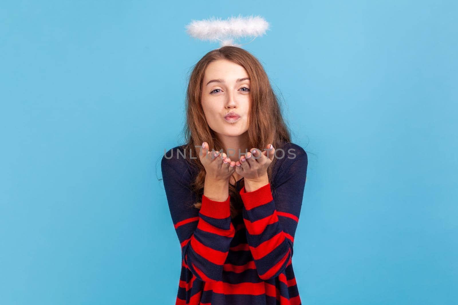 Kind woman wearing striped casual style sweater and nimb over head, sending air kiss over palms, expressing fondness, romantic feelings. Indoor studio shot isolated on blue background.