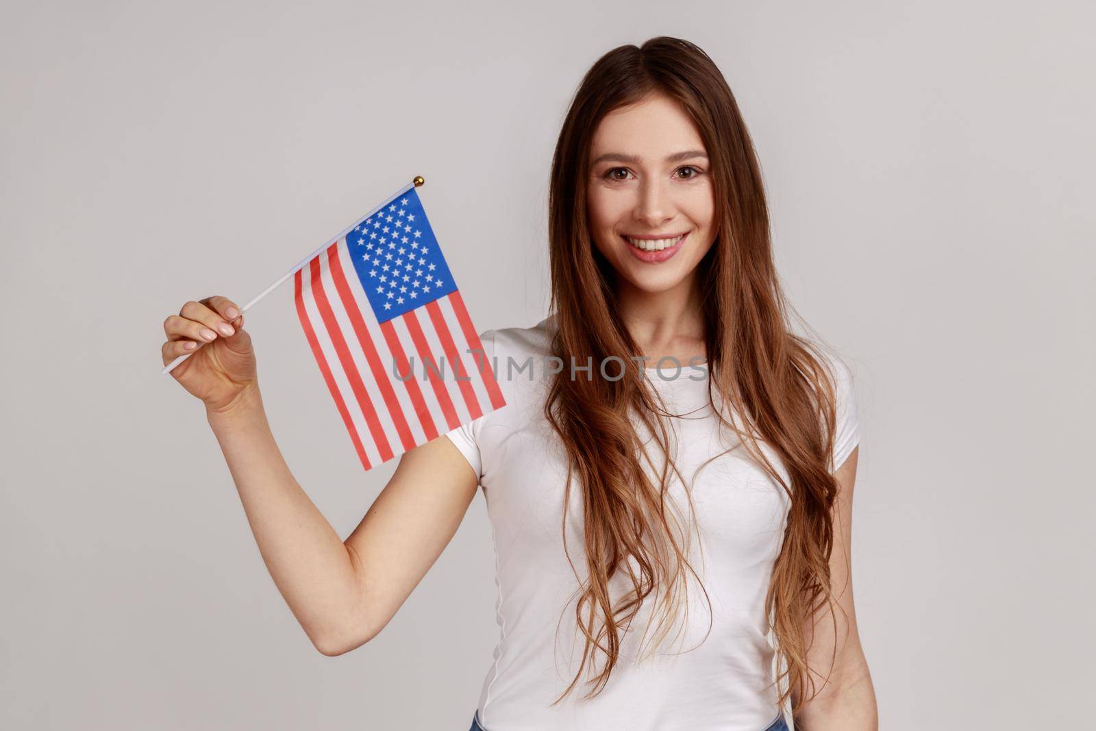 Portrait of smiling young adult woman holding USA national flag, celebrating national Independence Day - 4th july, wearing white T-shirt. Indoor studio shot isolated on gray background.