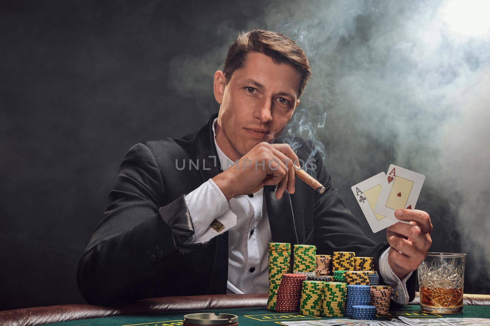 Elegant person in a black slassic suit and white shirt is playing poker sitting at the table at casino in smoke, against a white spotlight. He rejoicing his win showing two aces in his hand, smoking a cigar and looking at the camera. Gambling addiction. Sincere emotions and entertainment concept.