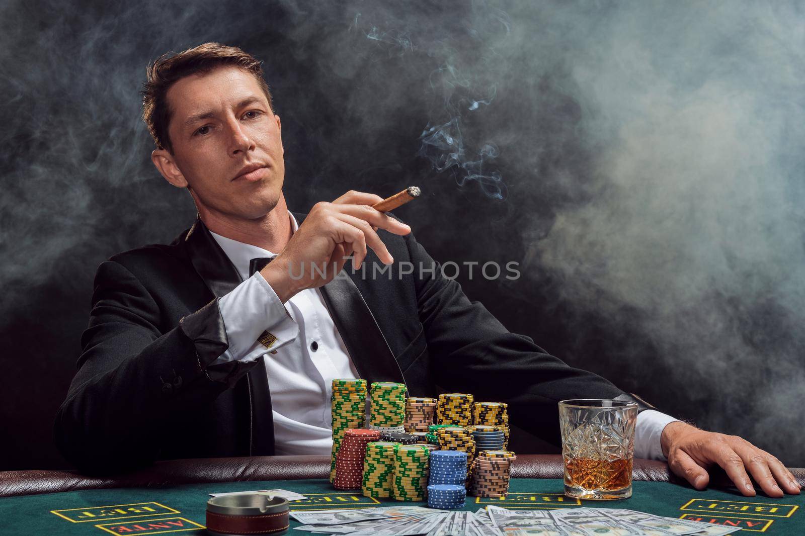 Smart person in a black slassic suit and white shirt is playing poker sitting at the table at casino in smoke, against a white spotlight. He rejoicing his victory smoking a cigar and looking at the camera. Gambling addiction. Sincere emotions and entertainment concept.