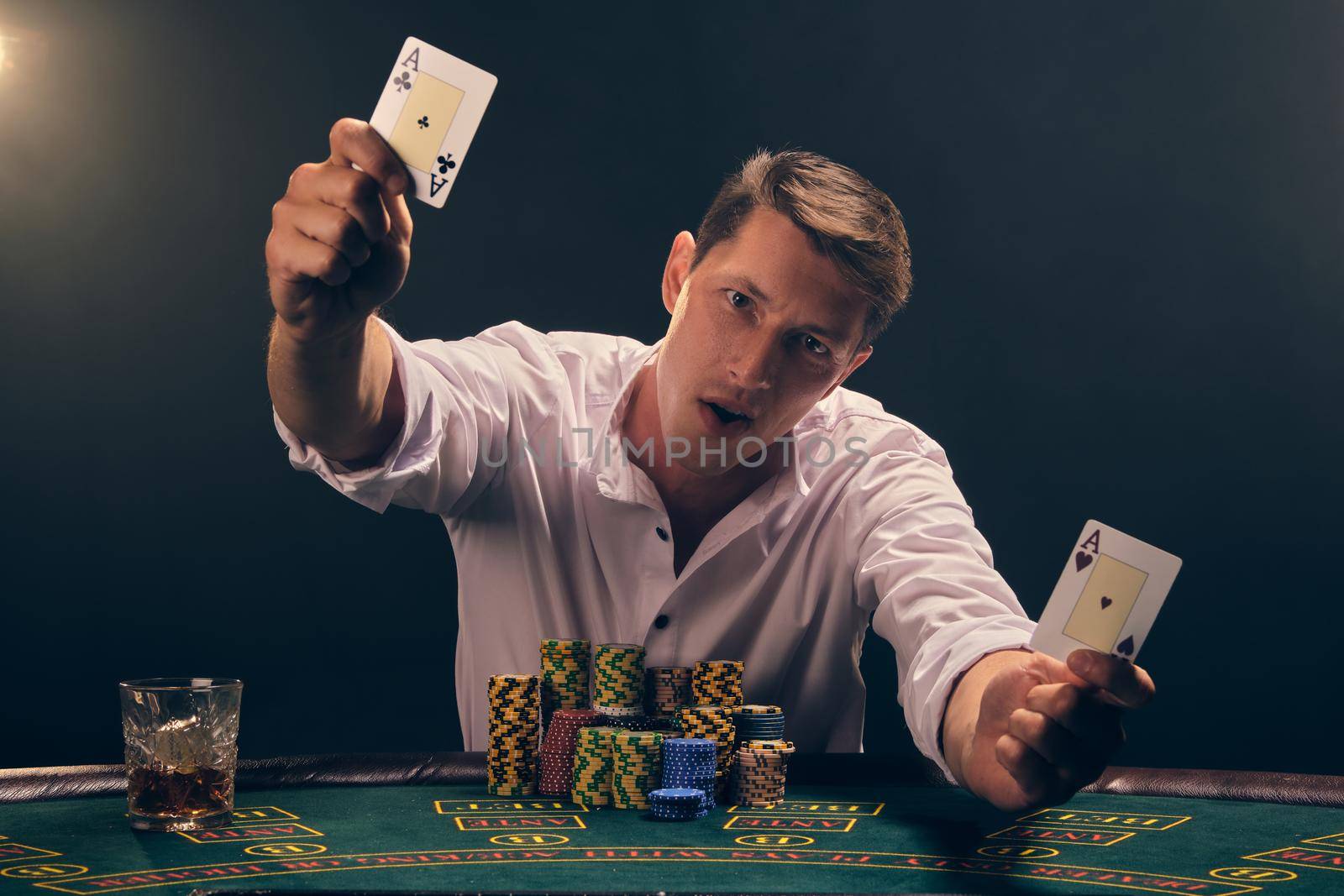 Handsome guy in a white shirt is playing poker sitting at the table at casino in smoke, against a white spotlight. He rejoicing his victory showing two aces in his hands and looking at the camera. Gambling addiction. Sincere emotions and entertainment concept.