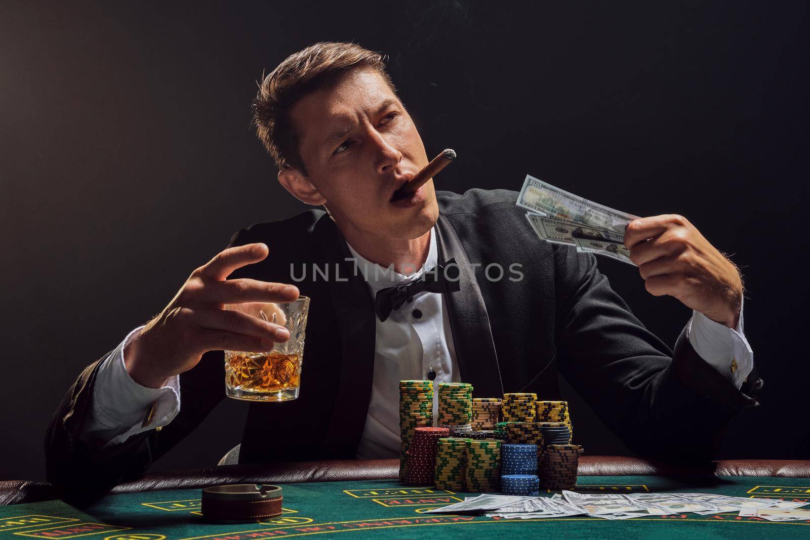 Handsome male in a black slassic suit and white shirt is playing poker sitting at the table at casino in smoke, against a white spotlight. He rejoicing his victory while smoking a cigar, drinking alcohol and holding some money in his hand. Gambling addiction. Sincere emotions and entertainment concept.