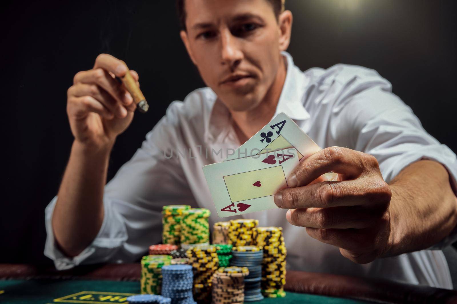Athletic guy in a white shirt is playing poker sitting at the table at casino in smoke, against a white spotlight. He rejoicing his victory and smoking a cigar while showing two aces in his hand. Gambling addiction. Sincere emotions and entertainment concept.