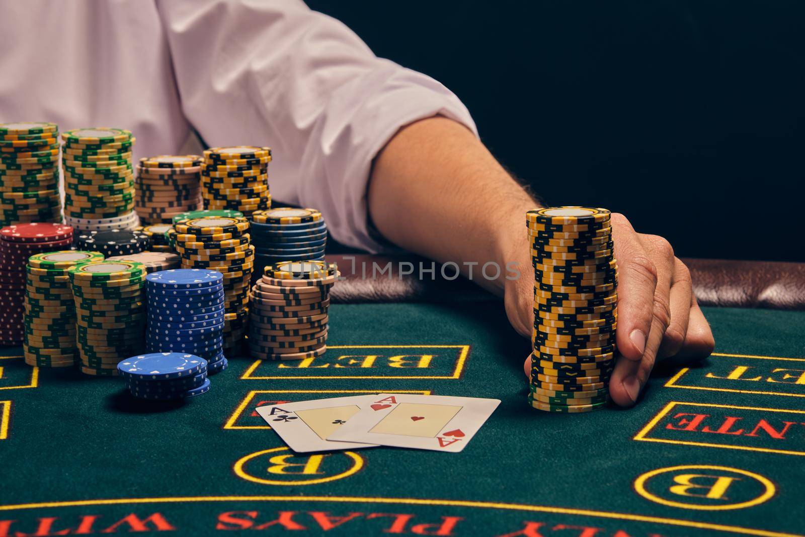 Close-up shot of an elegant fellow in a white shirt is playing poker sitting at the table at casino in smoke. He is holding some chips in his hand. Gambling addiction. Sincere emotions and entertainment concept.