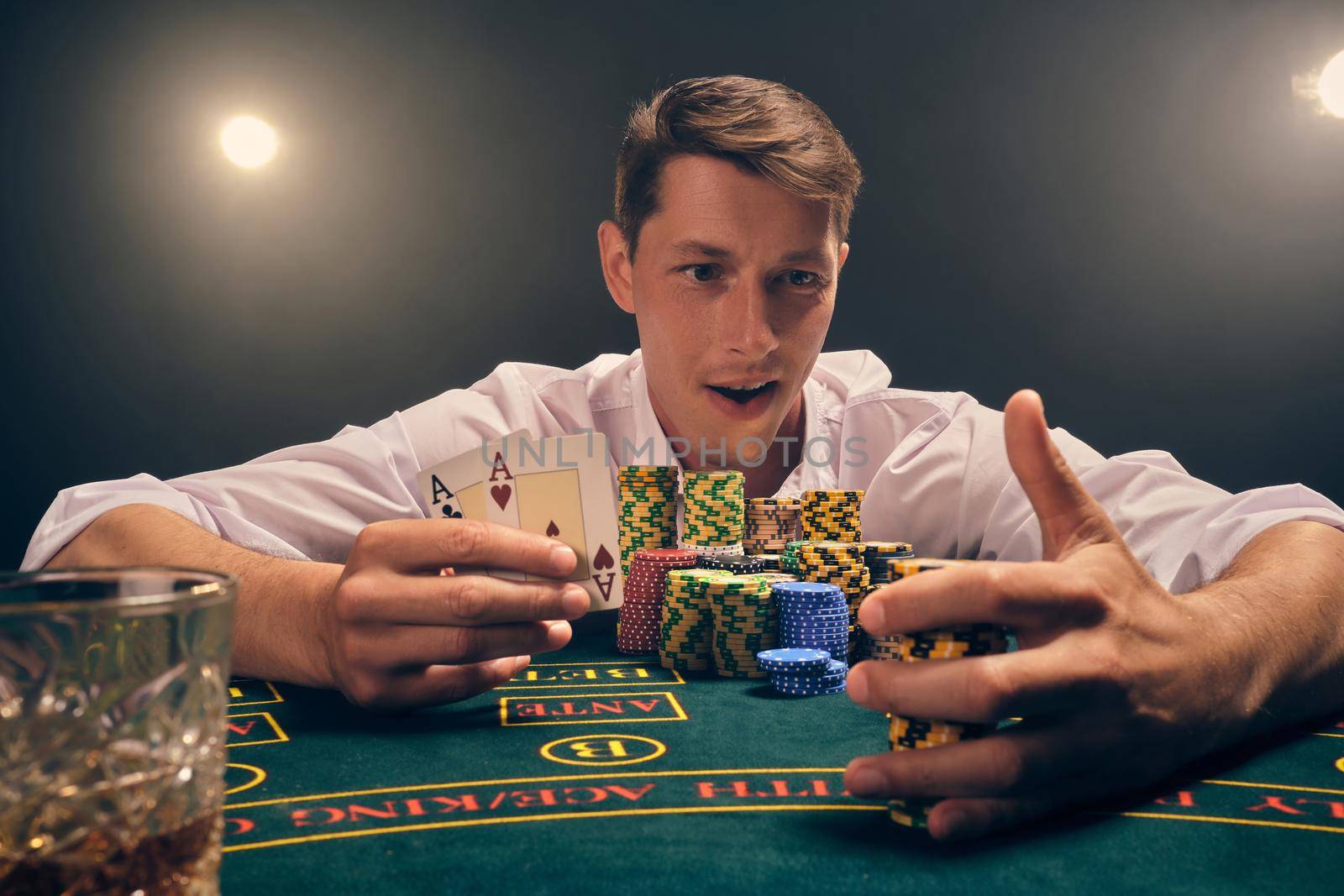 Close-up shot of a smart man in a white shirt is playing poker sitting at the table at casino in smoke, against a white spotlight. He rejoicing his victory showing two aces in his hand and holding some chips. Gambling addiction. Sincere emotions and entertainment concept.