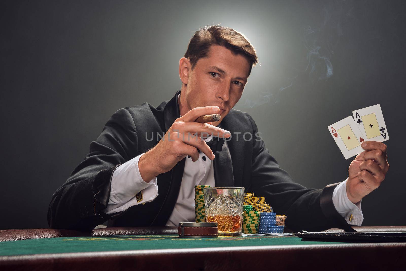 Stately guy in a black slassic suit and white shirt is playing poker sitting at the table at casino in smoke, against a white spotlight. He rejoicing his win showing two aces in his hand, smoking a cigar and looking at the camera. Gambling addiction. Sincere emotions and entertainment concept.