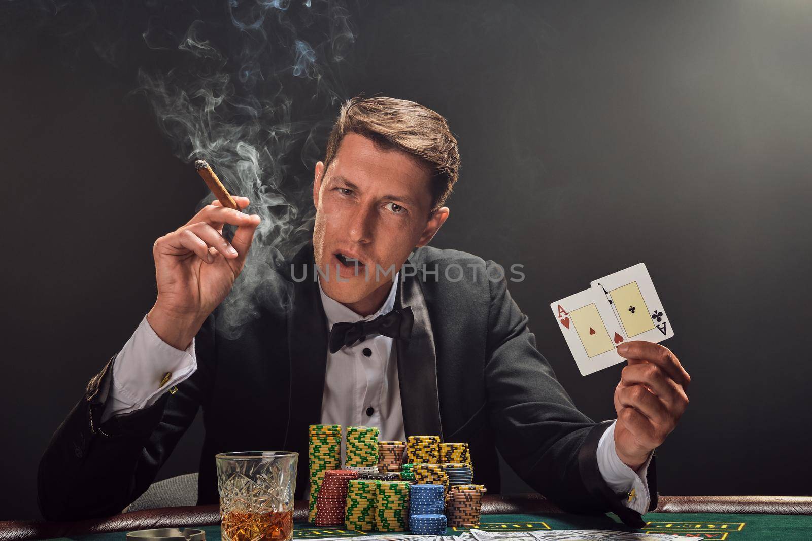 Good-looking guy in a black slassic suit and white shirt is playing poker sitting at the table at casino in smoke, against a white spotlight. He rejoicing his win showing two aces in his hand, smoking a cigar and looking at the camera. Gambling addiction. Sincere emotions and entertainment concept.
