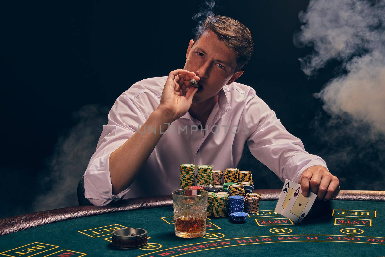 Stately male in a white shirt is playing poker sitting at the table at casino in smoke, against a white spotlight. He rejoicing his victory and smoking a cigar while looking at the camera. Gambling addiction. Sincere emotions and entertainment concept.