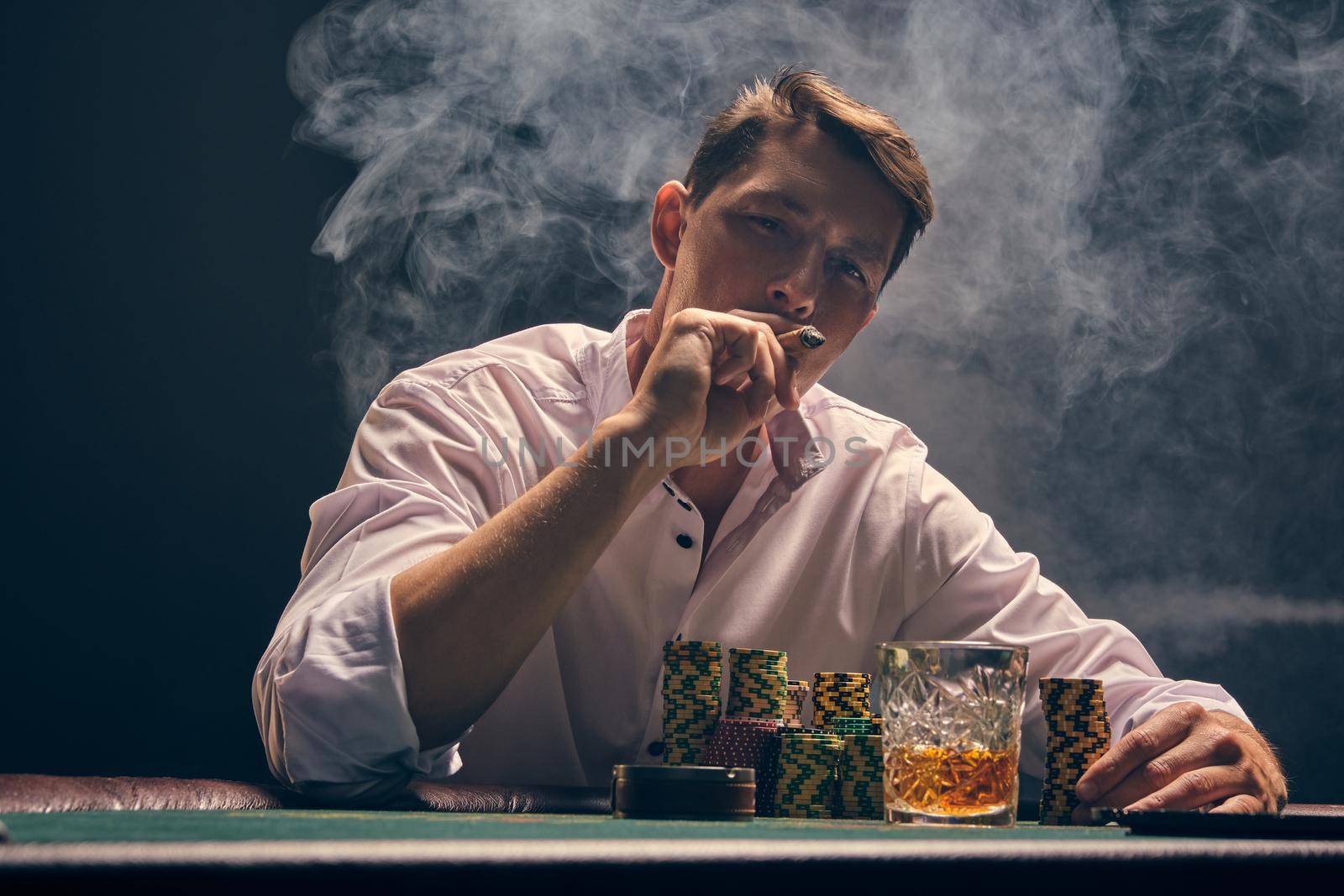 Handsome person in a white shirt is playing poker sitting at the table at casino in smoke, against a white spotlight. He rejoicing his victory and smoking a cigar. Gambling addiction. Sincere emotions and entertainment concept.