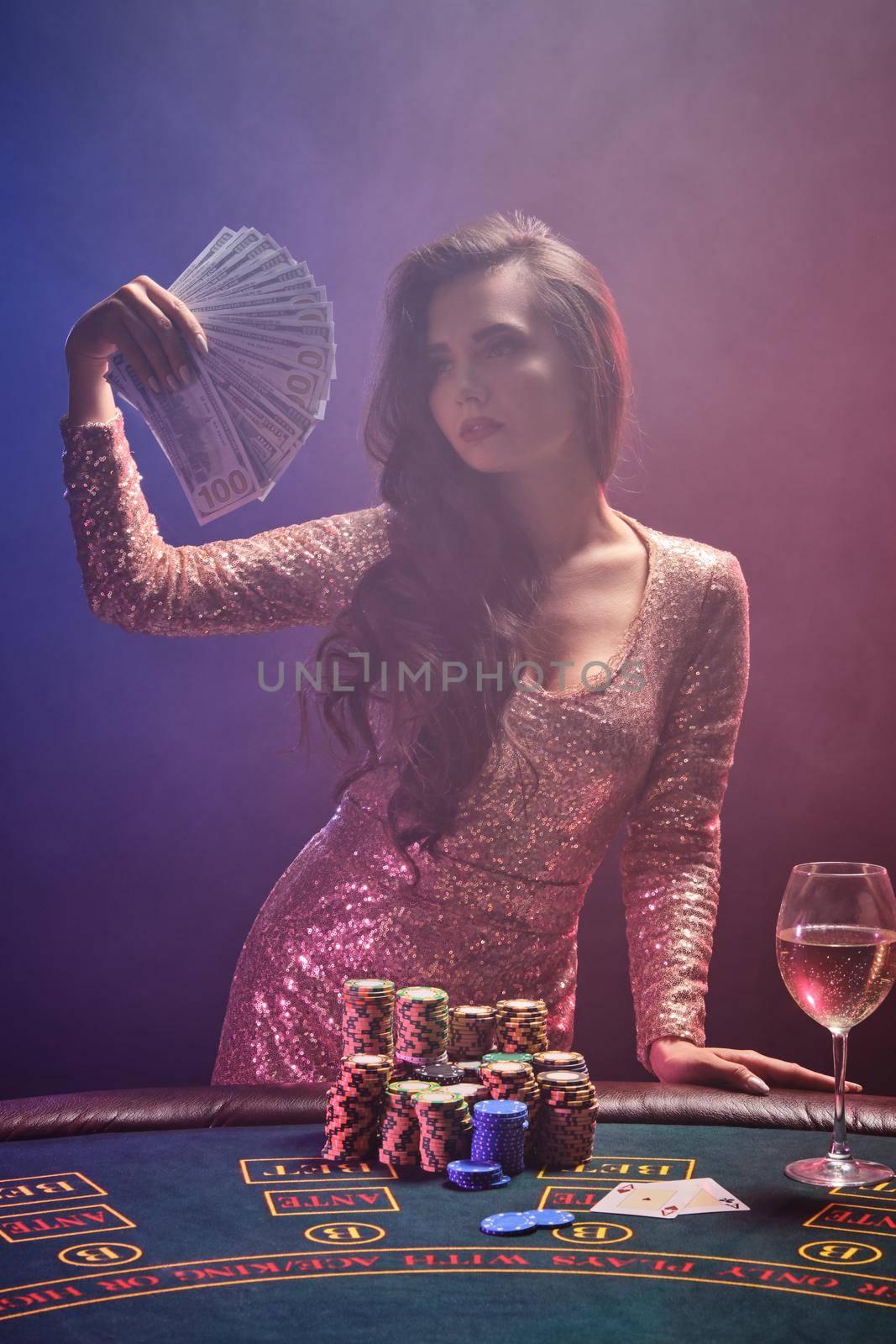 Portrait of a sexy brunette lady with bright make-up, dressed in a golden glittered dress, posing against a gambling table with a fan of hundred dollar bills in her hands and looking at it. Poker concept on a black smoke background with pink and blue backlights. Casino.