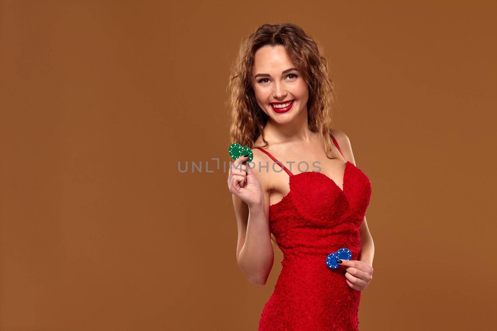 Attractive young caucasian brown-haired woman in red cocktail dress on brown background holding few poker chips. Casino concept, gambling industry