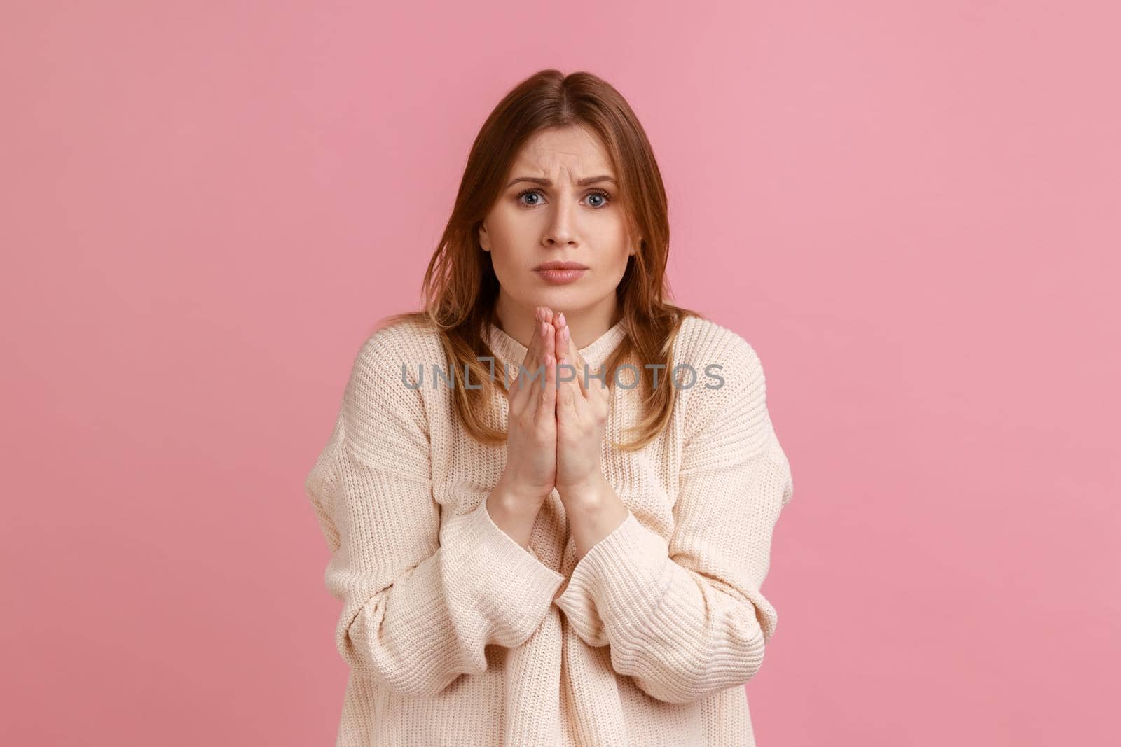 Portrait of attractive blond woman praying, begging with naughty and pleading grimace, gesture of asking apologizing, wearing white sweater. Indoor studio shot isolated on pink background.