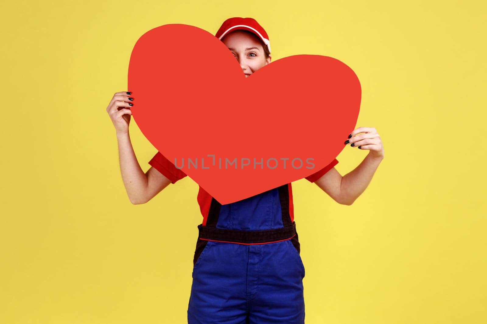 Portrait of optimistic worker woman peeping from big red heart, expressing romantic feelings, looking at camera, wearing overalls and red cap. Indoor studio shot isolated on yellow background.