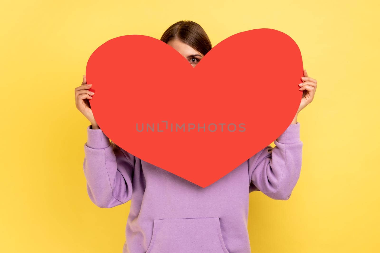 Positive woman hiding her face behind big red heart and looking at camera with curious prying eyes, feeling affection fondness, wearing purple hoodie. Indoor studio shot isolated on yellow background.