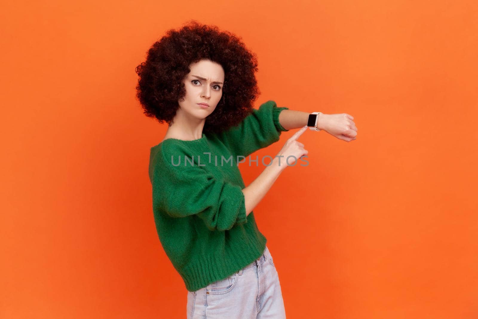 Bossy woman with Afro hairstyle wearing green casual style sweater pointing wrist watch and looking annoyed displeased, showing clock to hurry up. Indoor studio shot isolated on orange background.