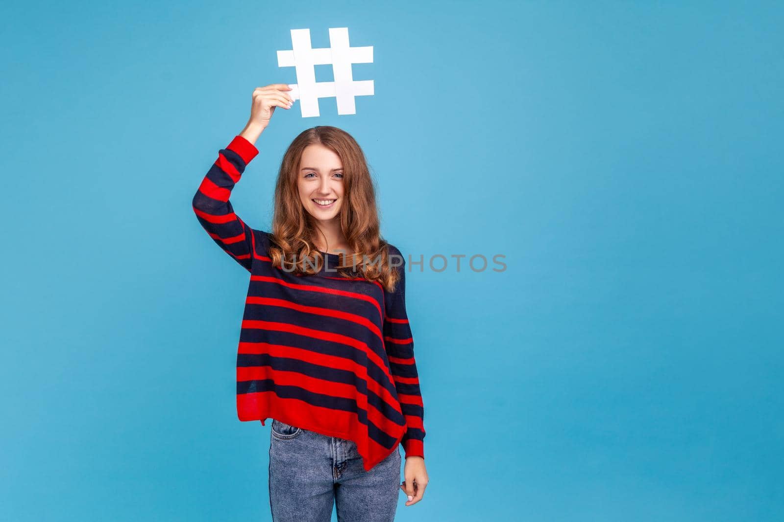 Portrait of woman wearing striped casual style sweater, standing holding hashtag over head, promoting viral topic, trendy idea, smiling to camera. Indoor studio shot isolated on blue background.