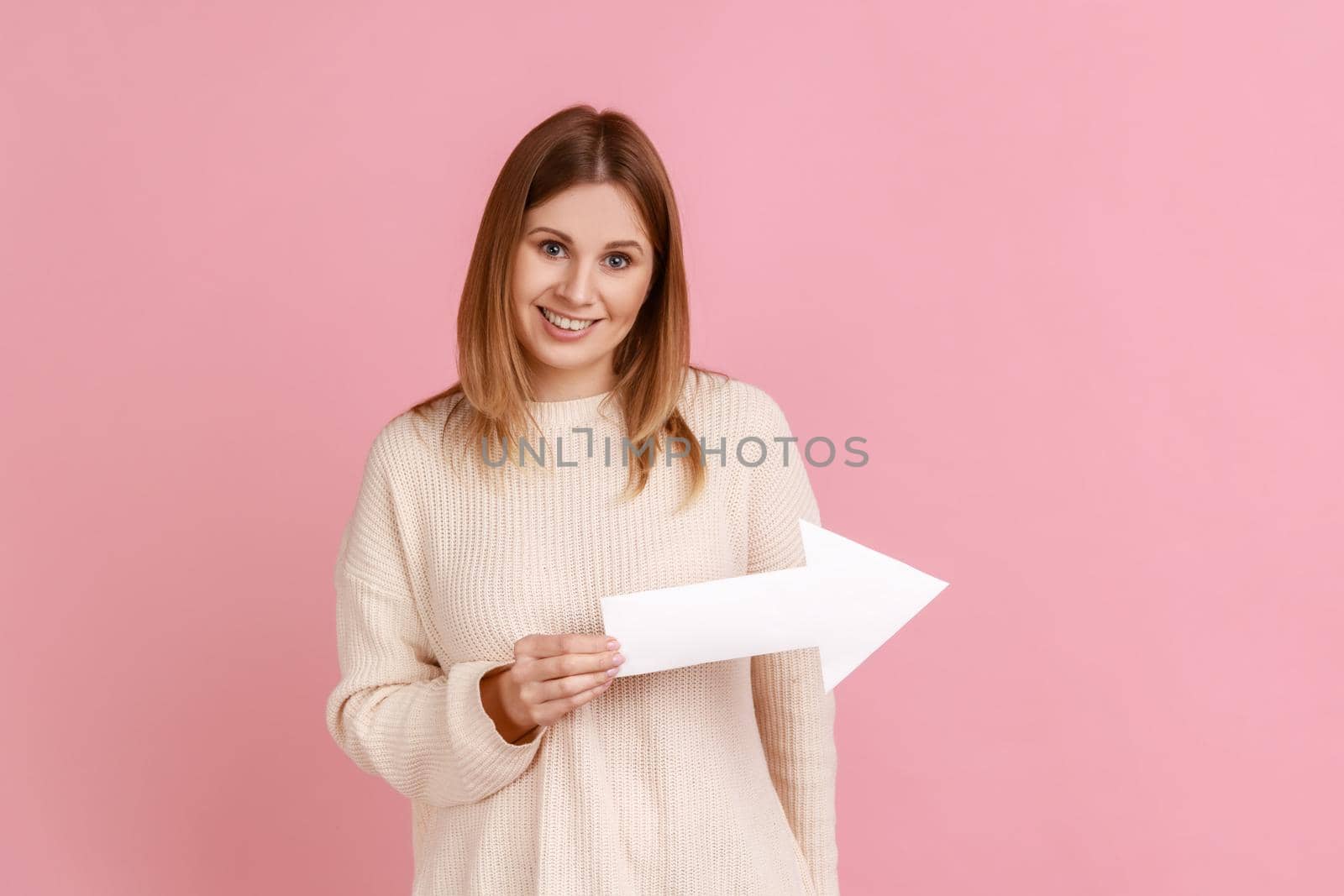 Portrait of smiling satisfied blond woman pointing direction with arrow aside, looking at camera with positive expression, wearing white sweater. Indoor studio shot isolated on pink background.