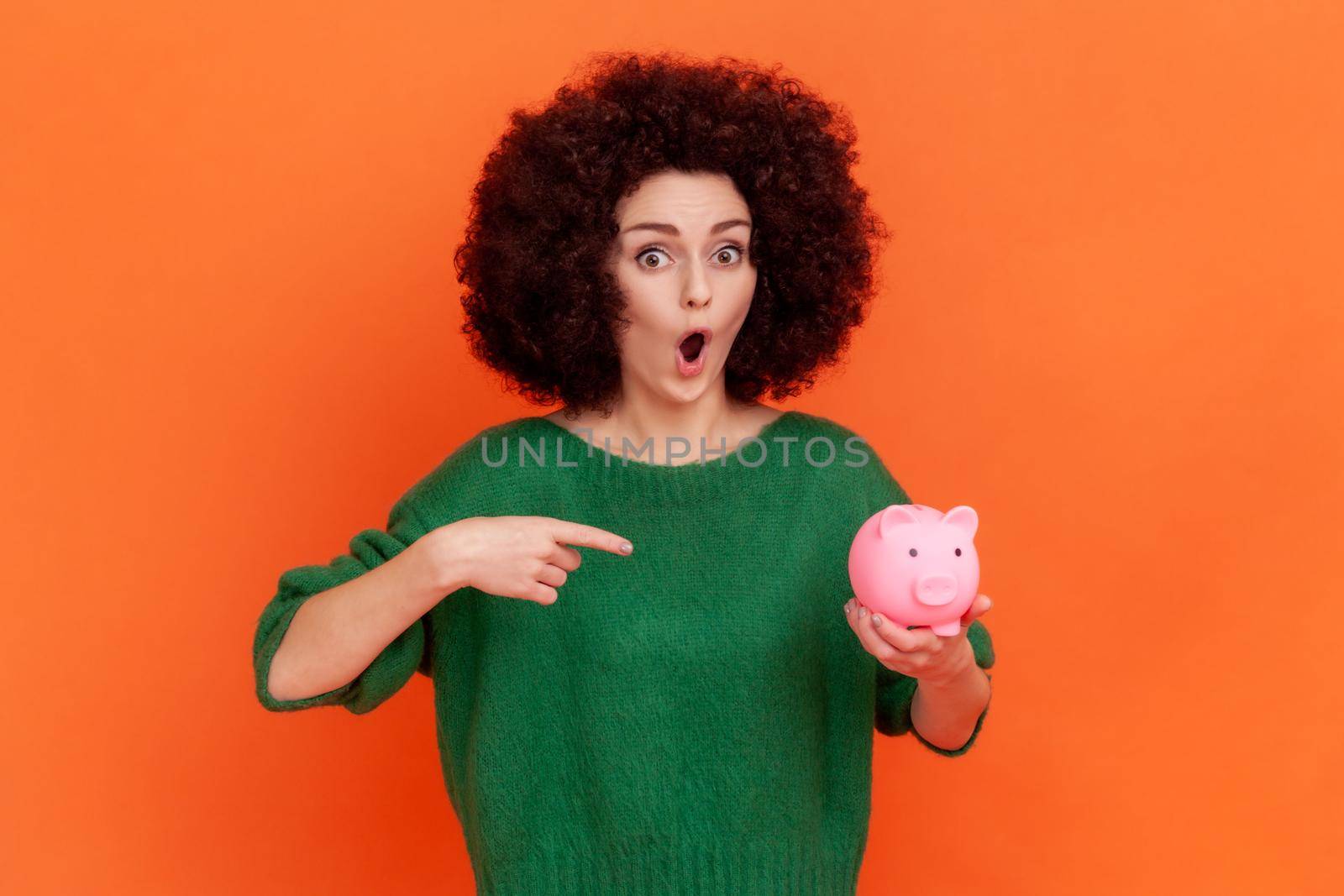 Portrait of shocked woman with Afro hairstyle wearing green casual style sweater standing pointing at piggy bank in her hand, advantageous bank offer. Indoor studio shot isolated on orange background.