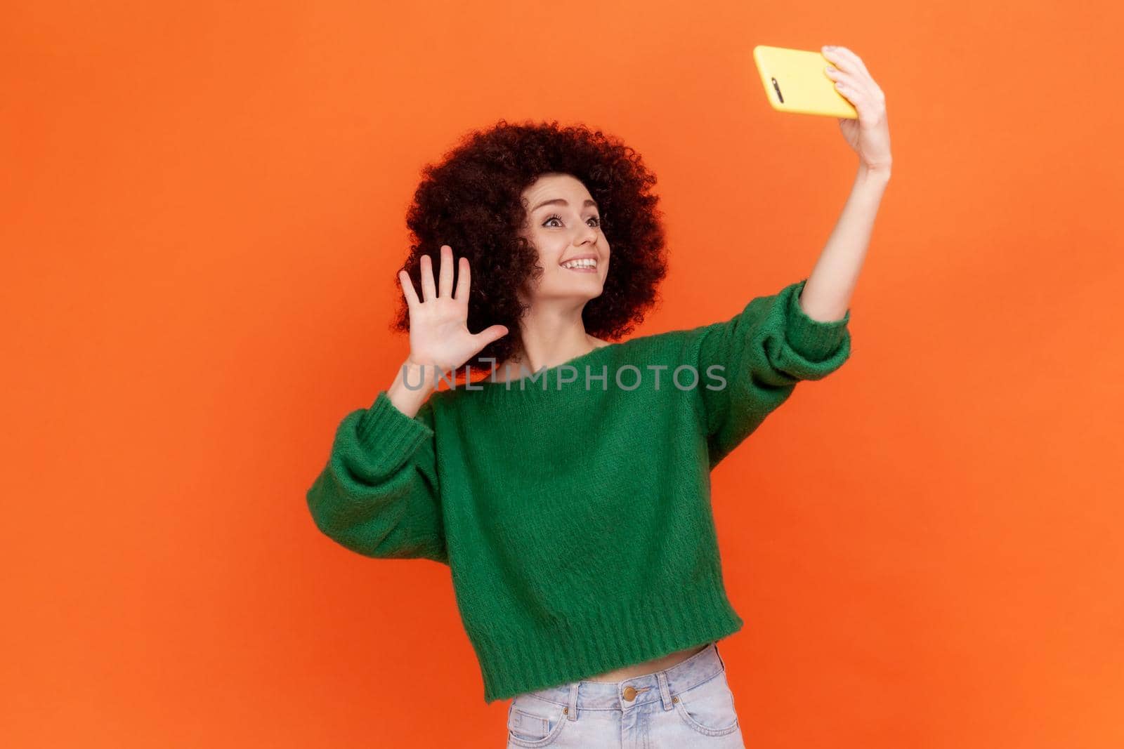 Adorable blogger woman with Afro hairstyle wearing green casual style sweater waving hand to followers while having livestream, greeting new people. Indoor studio shot isolated on orange background.