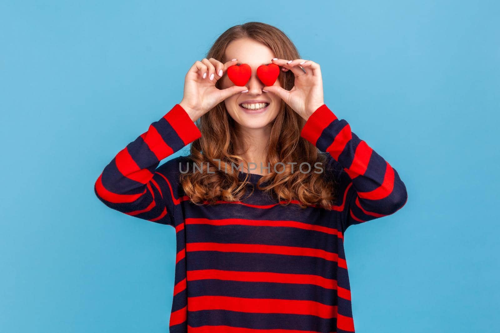 Funny positive woman wearing striped casual style sweater, covering eyes with small red hearts, love symbol, smiling toothily, falling in love. Indoor studio shot isolated on blue background.