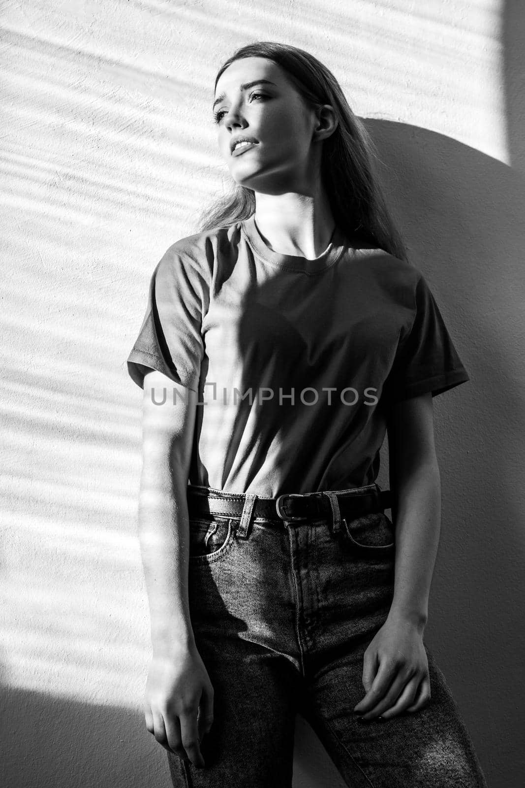 Young adult beautiful woman wearing T-shirt and jeans standing looking away with pensive romantic facial expression. Black and white photography, indoor studio shot illuminated by sunlight from window