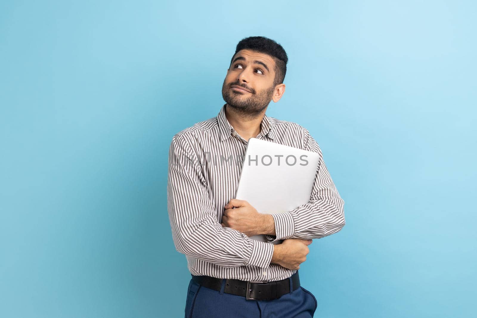 Portrait of pensive businessman standing holding closed laptop or folder, looking away with thoughtful expression, wearing striped shirt. Indoor studio shot isolated on blue background.
