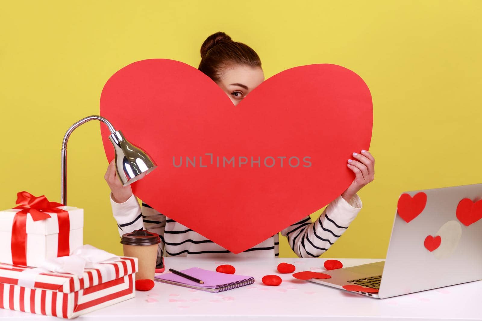 Portrait of shy woman sitting at workplace, holding and peeping from big red heart, expressing romantic feelings and love. Indoor studio studio shot isolated on yellow background.