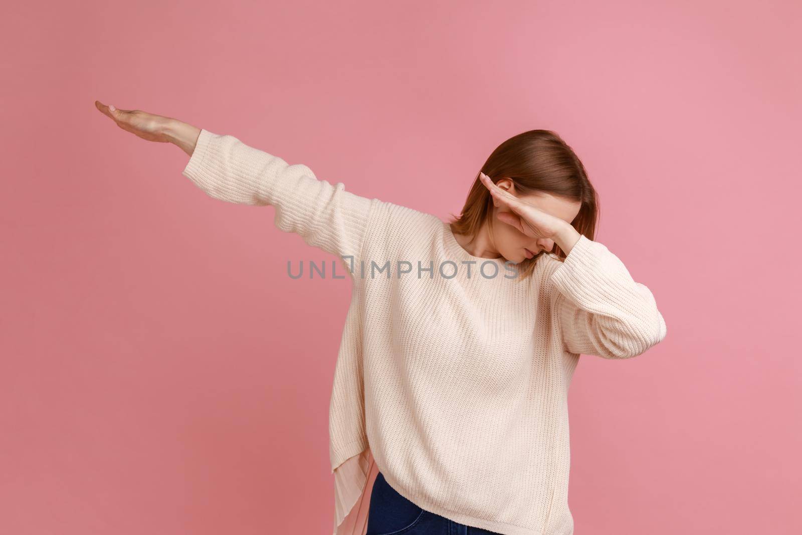Portrait of blond woman showing dab dance pose, famous internet meme of triumph, performing dabbing trends with hand gesture, wearing white sweater. Indoor studio shot isolated on pink background.