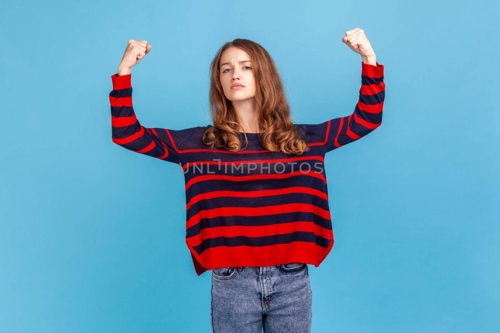 Portrait of confident independent woman, showing biceps, feeling energy, confidence and power in hands to win, become successful, wearing sweater. Indoor studio shot isolated on blue background.