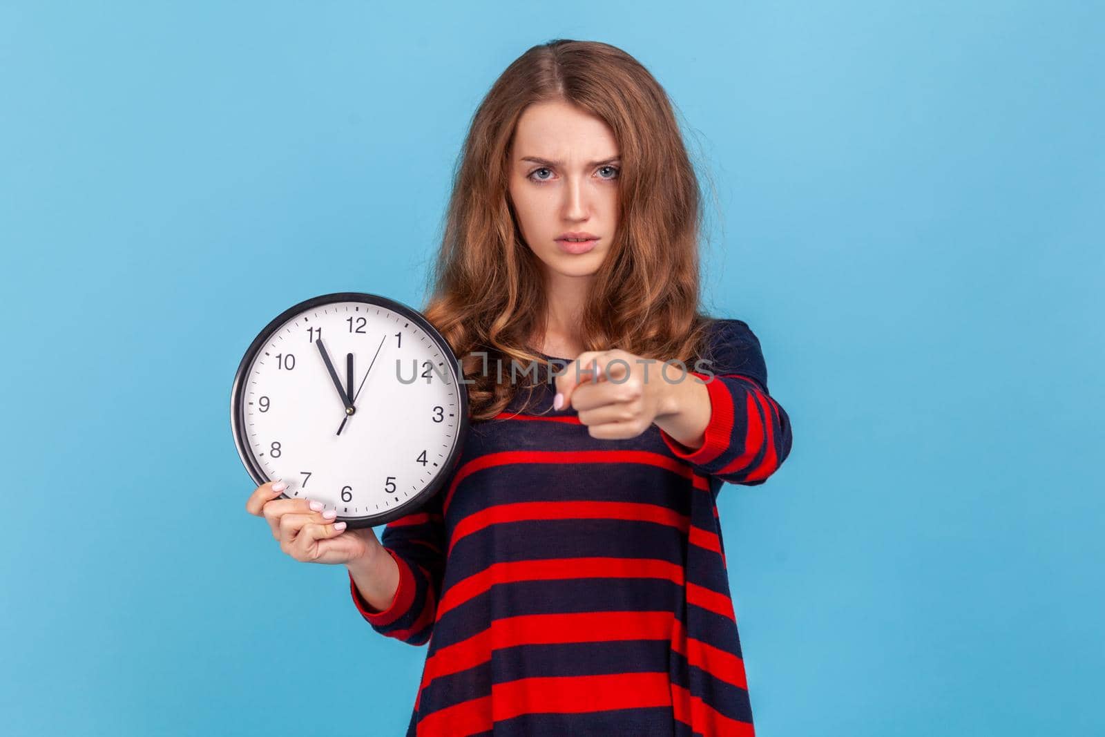 Portrait of serious bossy woman wearing striped casual style sweater, pointing finger at you holding in hand big wall clock, motivation. Indoor studio shot isolated on blue background.