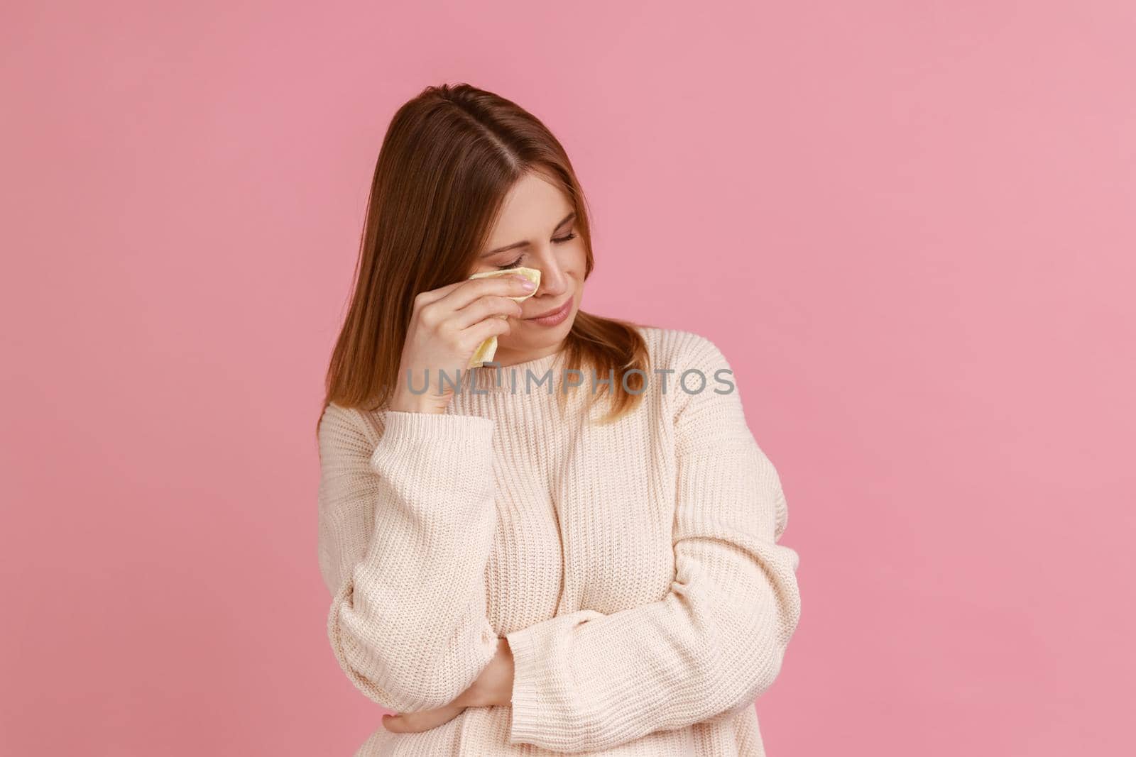 Portrait of blond woman frowning, wiping tears crying, feeling desperate hopeless, coping lonely in emotional stress, wearing white sweater. Indoor studio shot isolated on pink background.