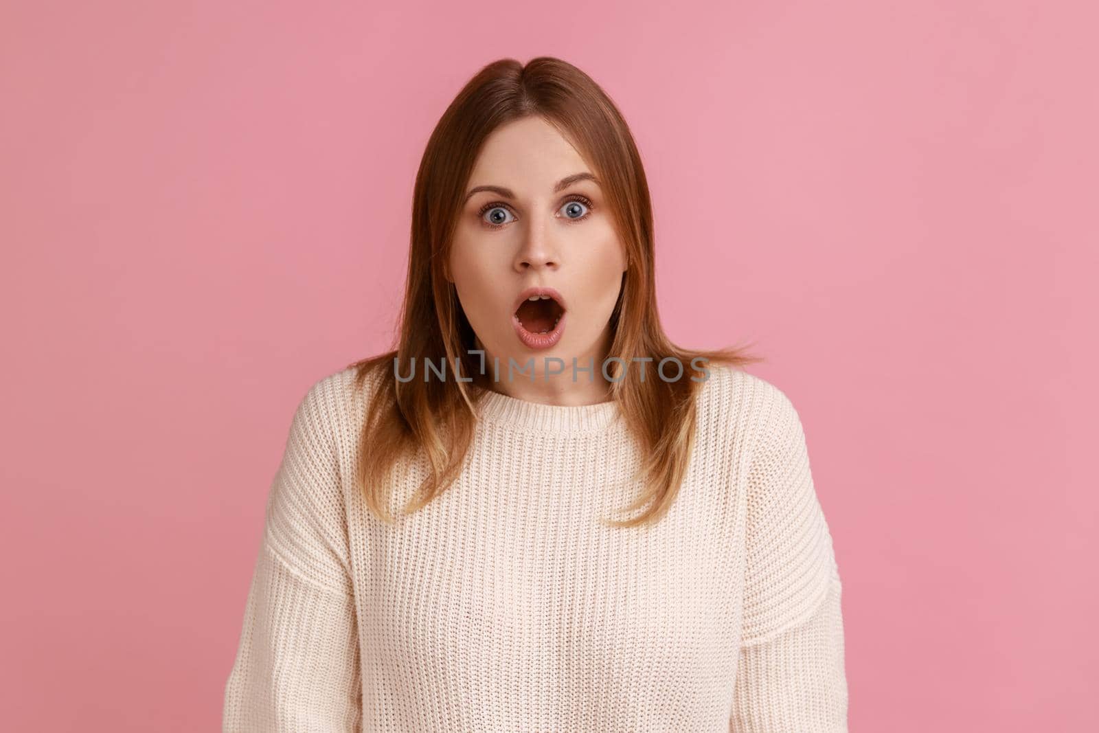 Portrait of shocked blond woman standing looking at camera with open mouth, being astonished by breaking news, wearing white sweater. Indoor studio shot isolated on pink background.