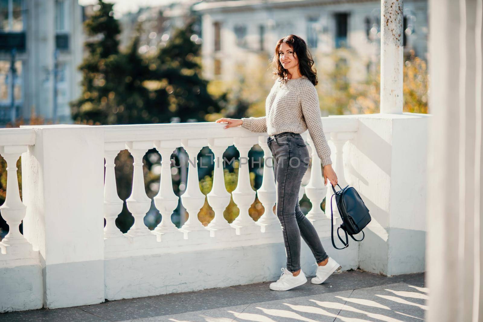 Lifestyle young woman in jeans, white sneakers, beige sweater. She is holding a backpack in one hand. Against the background of autumn trees and white balusters